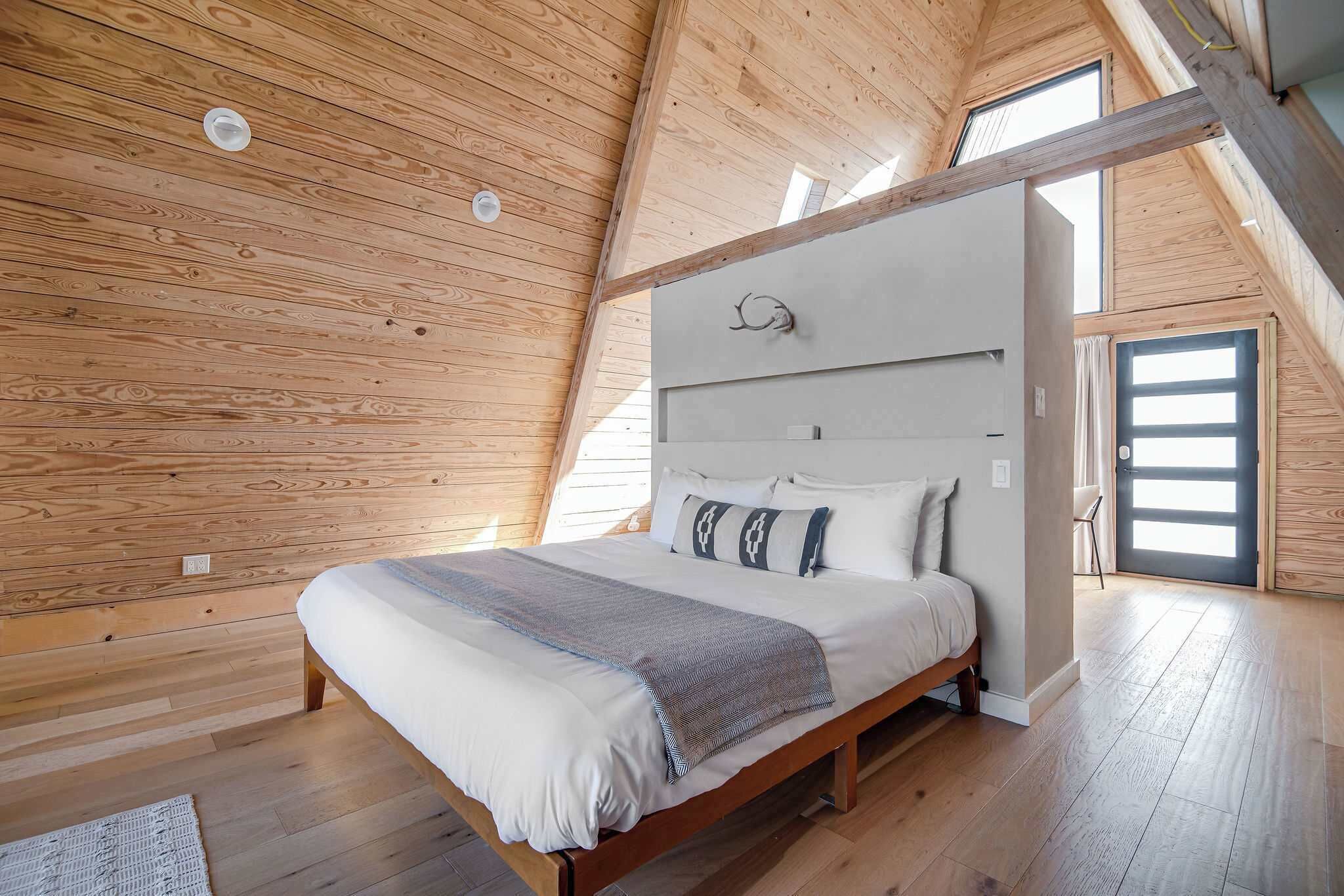 A high-ceilinged bedroom with a wooden ceiling, a bed with a white mattress and gray blanket, and a large window.