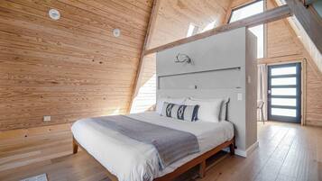 A high-ceilinged bedroom with a wooden ceiling, a bed with a white mattress and gray blanket, and a large window.
