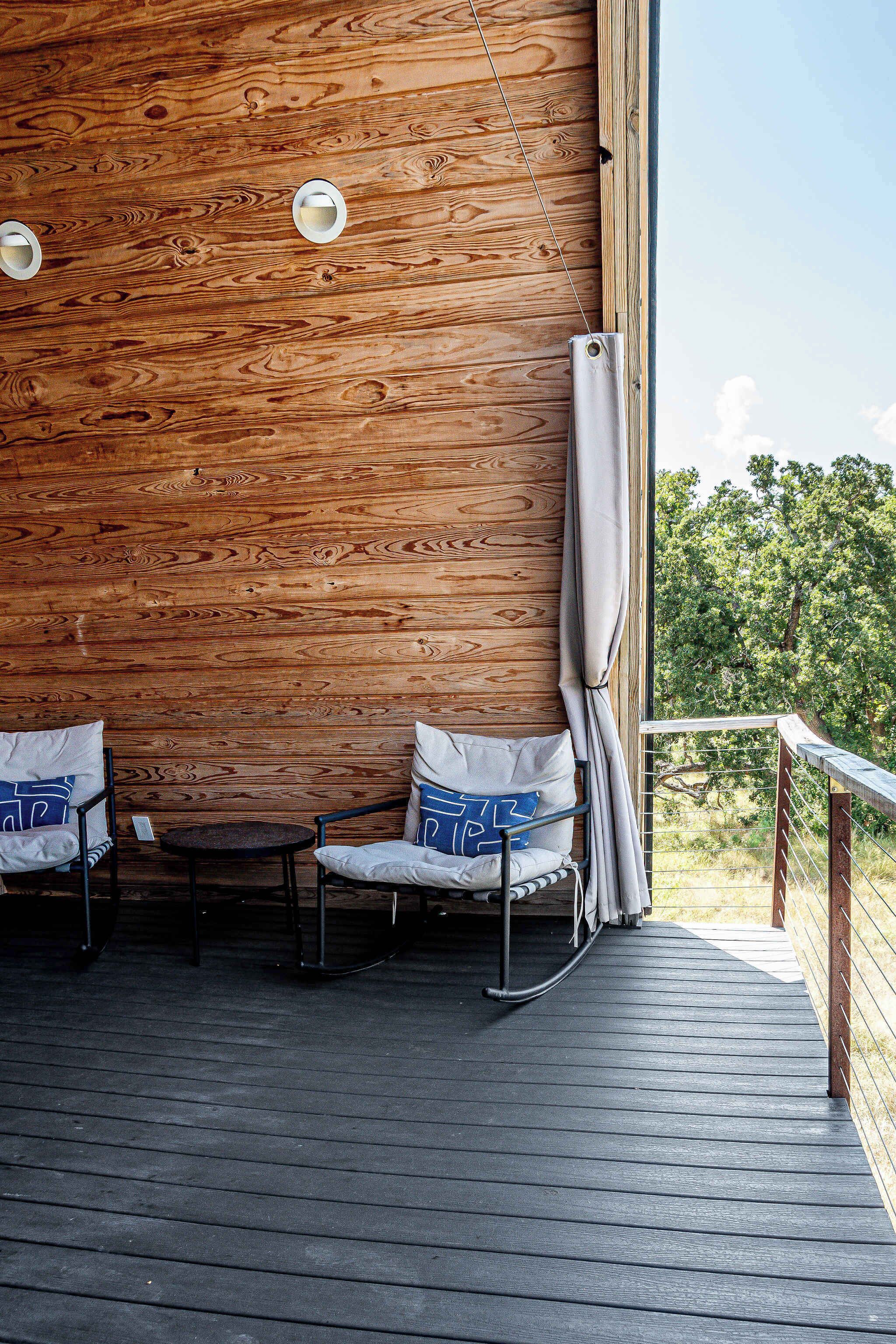 A wooden deck with two chairs and a small table, a wooden wall, and a view of trees.