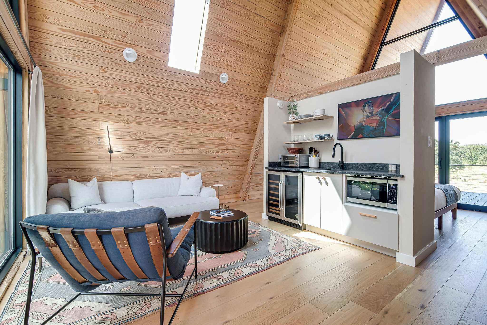 A modern loft with a wooden ceiling, a white sofa, a blue chair, a kitchen with a microwave, and a large window.