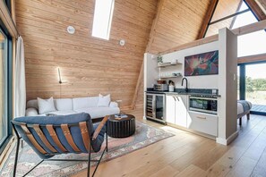 A modern loft with a wooden ceiling, a white sofa, a blue chair, a kitchen with a microwave, and a large window. - A-Frame Ranch: Mesquite Treehouse with Shared Pool (Fredericksburg)