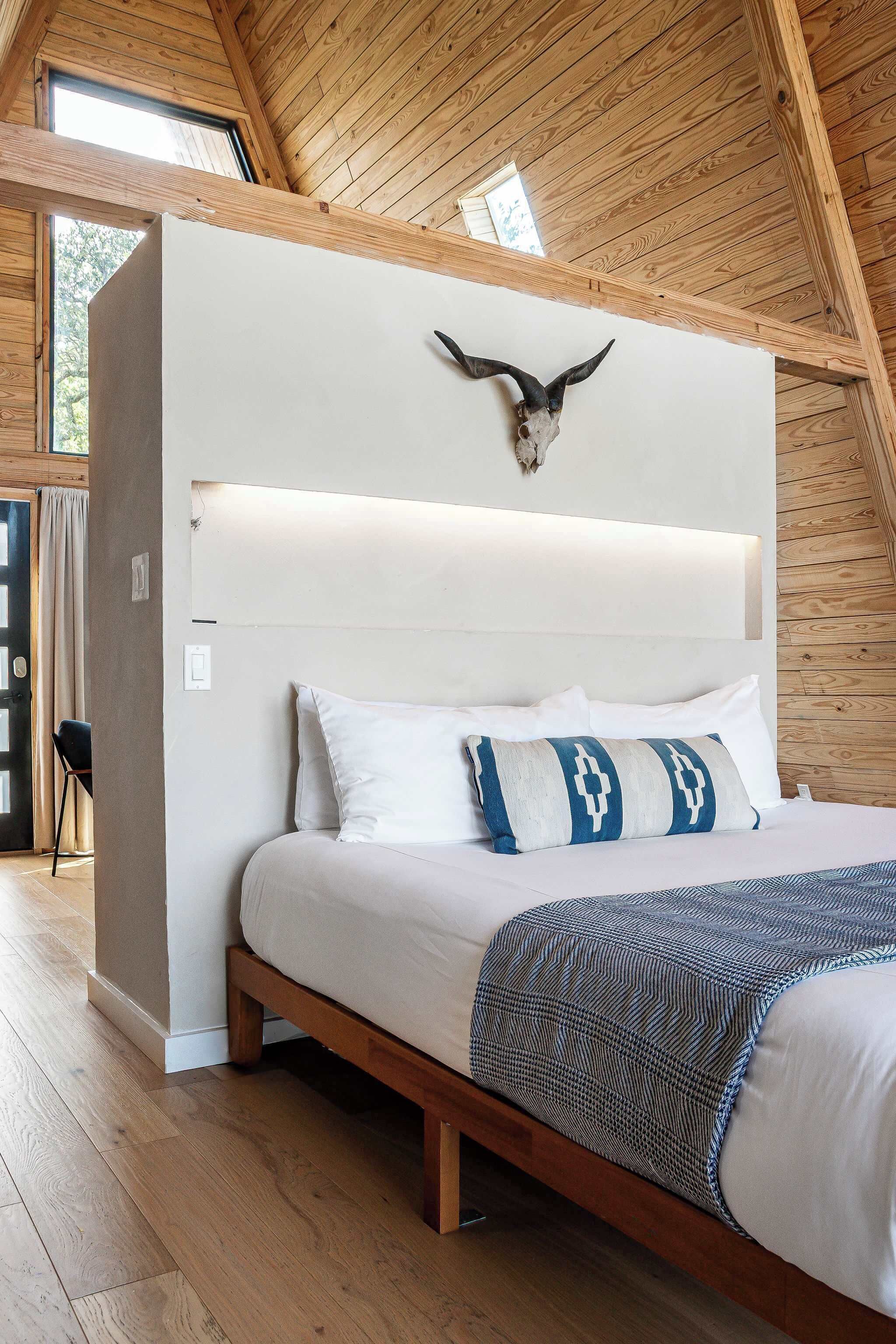 A bedroom with a wooden ceiling, a bed with white bedding and patterned pillows, and a wall-mounted headboard.