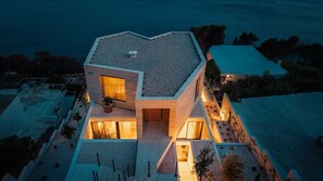 A modern house with a unique geometric roof, a visible entrance, and a well-lit interior. - Villa Aurea Beachfront & Spa with Heated Pool (Mimice)