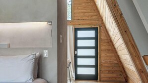 A room with a sloped ceiling, wooden beams, a bed, a door, and a view through a window. - A-Frame Ranch: Pecan Treehouse with Shared Pool (Fredericksburg)