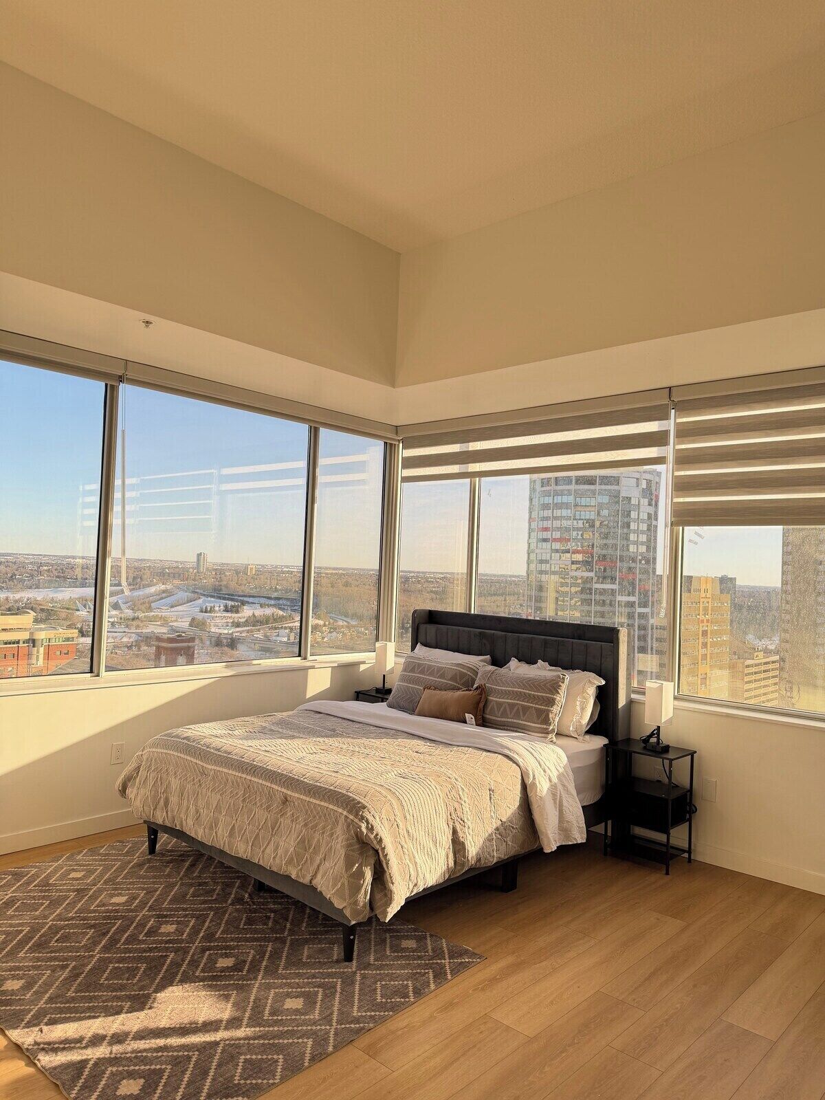 A bedroom with a large bed, a nightstand, and a view of a cityscape through large windows.