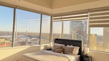A bedroom with a large bed, a nightstand, and a view of a cityscape through large windows.