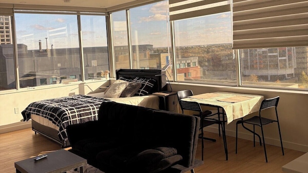 A modern bedroom with a large bed, a desk, and a chair. There are large windows with blinds, and the room has a high ceiling.