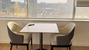 A modern room with a table and two chairs, large windows with blinds, and a view of a cityscape.