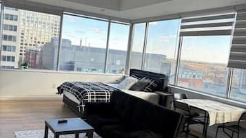 A spacious living room with a large bed, a black sofa, a small table, and a dining area with a table and chairs.