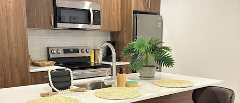 Una cocina moderna con armarios de madera, electrodomésticos de acero inoxidable y encimera blanca. Hay tres taburetes de bar con asientos negros y bases metálicas.