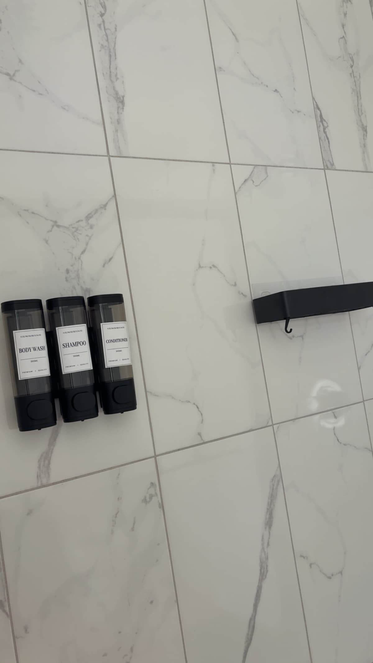 Three toiletry bottles labeled body wash, shampoo, and conditioner on a tiled floor.