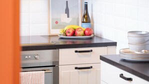 Une cuisine avec des meubles blancs, un tableau encadré, une bouteille de vin et une assiette de fruits.