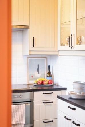 A kitchen with white cabinets, a framed picture, a bottle of wine, and a plate of fruit. - La Petite Crevette: Beach and Charm in De Haan (De Haan)