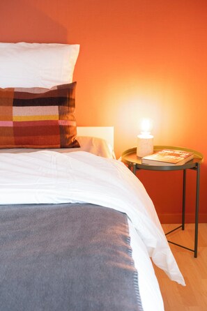 Una cama bien hecha con edredón blanco y gris, un cojín colorido y una mesita de noche con una lámpara y un libro.