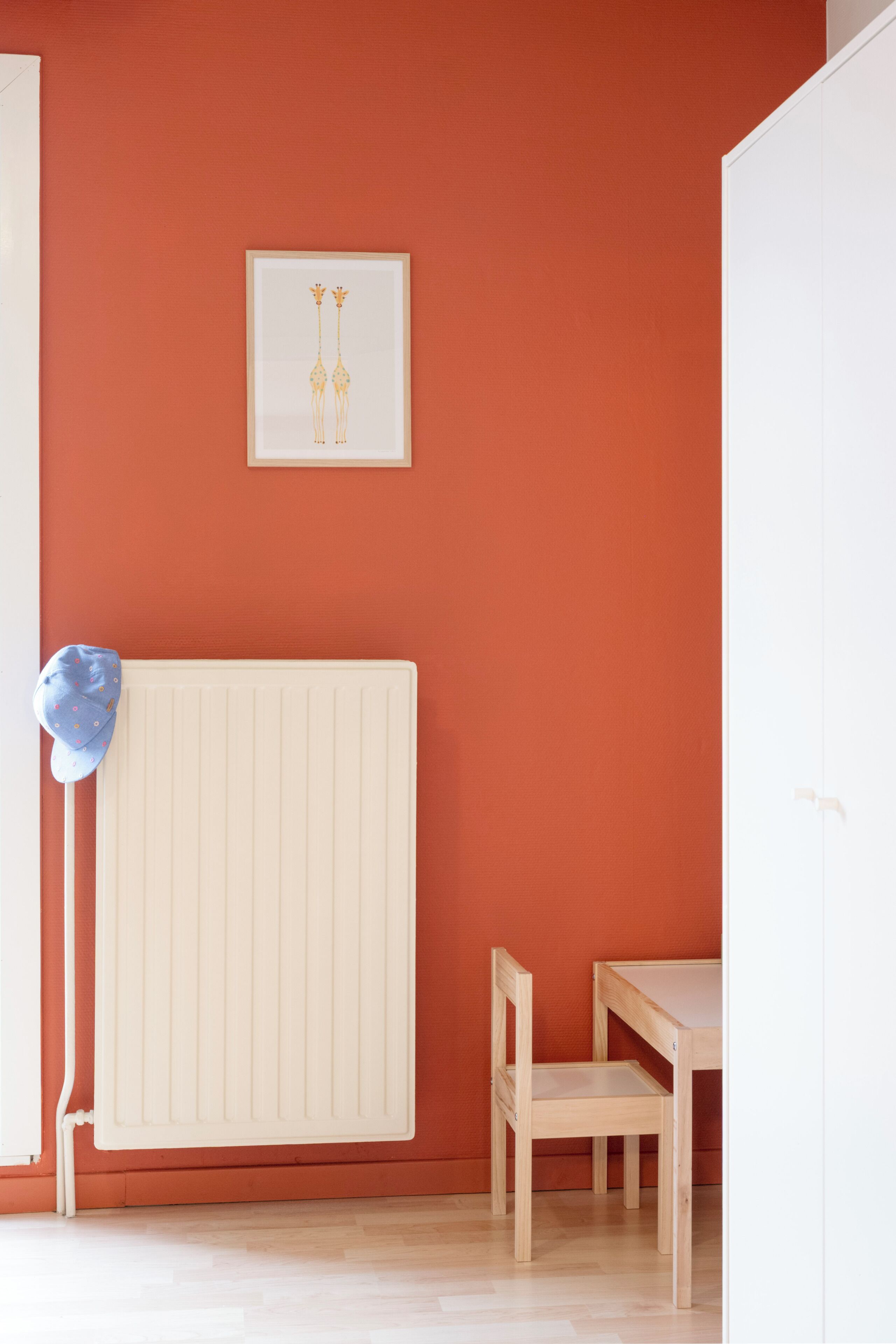 Una habitación con un radiador, un cuadro en la pared, una gorra azul sobre el radiador y una mesa de madera con una silla.