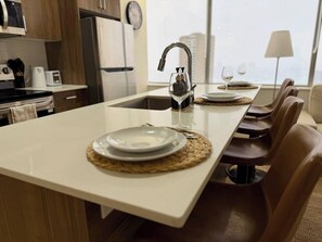 A modern kitchen with a central island, a built-in sink, and a dining area with chairs and a table set for two. - Downtown Gem 2BR Apartment with Stunning City View (Edmonton)