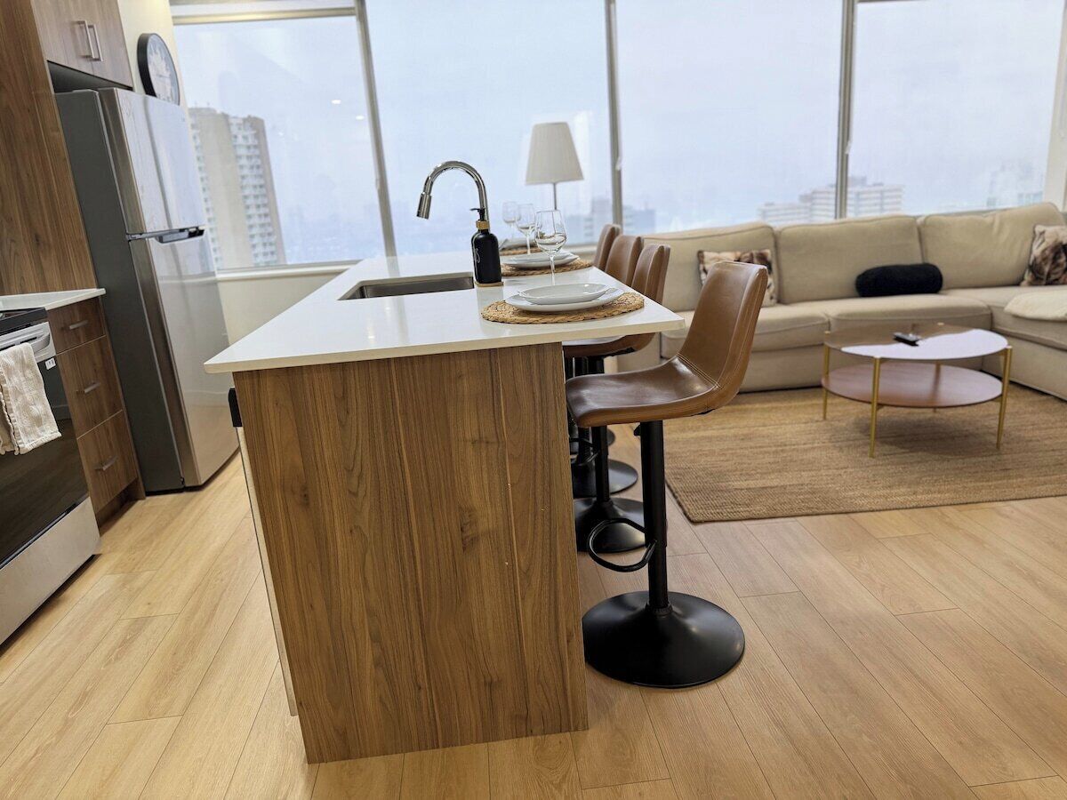 Una cocina moderna con una isla central, taburetes de bar, un área de comedor con sofá y vistas a la ciudad a través de amplios ventanales.