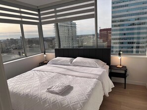 A hotel room with a large bed, a nightstand, a lamp, and a view of the city through large windows. - Downtown Gem 2BR Apartment with Stunning City View (Edmonton)
