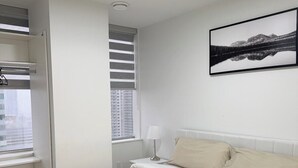 Ein Schlafzimmer mit Bett, Fenster mit Jalousien, Nachttisch mit Lampe und einem gerahmten Bild an der Wand.