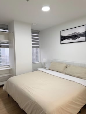 A bedroom with a bed, a window with blinds, a nightstand with a lamp, and a framed picture on the wall. - Downtown Gem 2BR Apartment with Stunning City View (Edmonton)