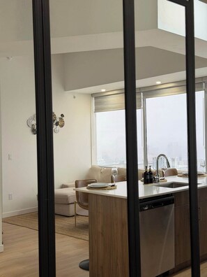 A modern kitchen with a dishwasher, a dining area with a sofa and table, and a large window with a view. - Downtown Gem 2BR Apartment with Stunning City View (Edmonton)