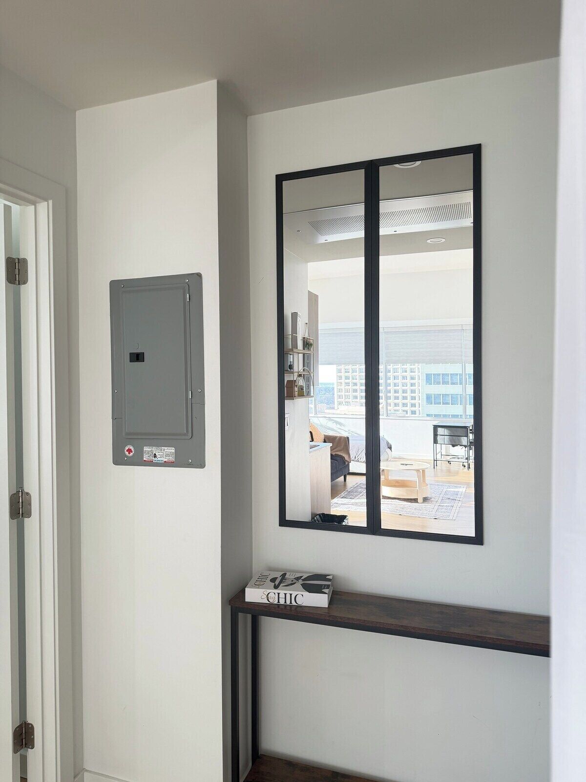 A room with a white wall, a door, a window with a view of a cityscape, and a shelf with a book.