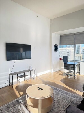 A modern living room with a flat-screen TV, a round wooden table, a sofa, and a view of a cityscape through large windows.