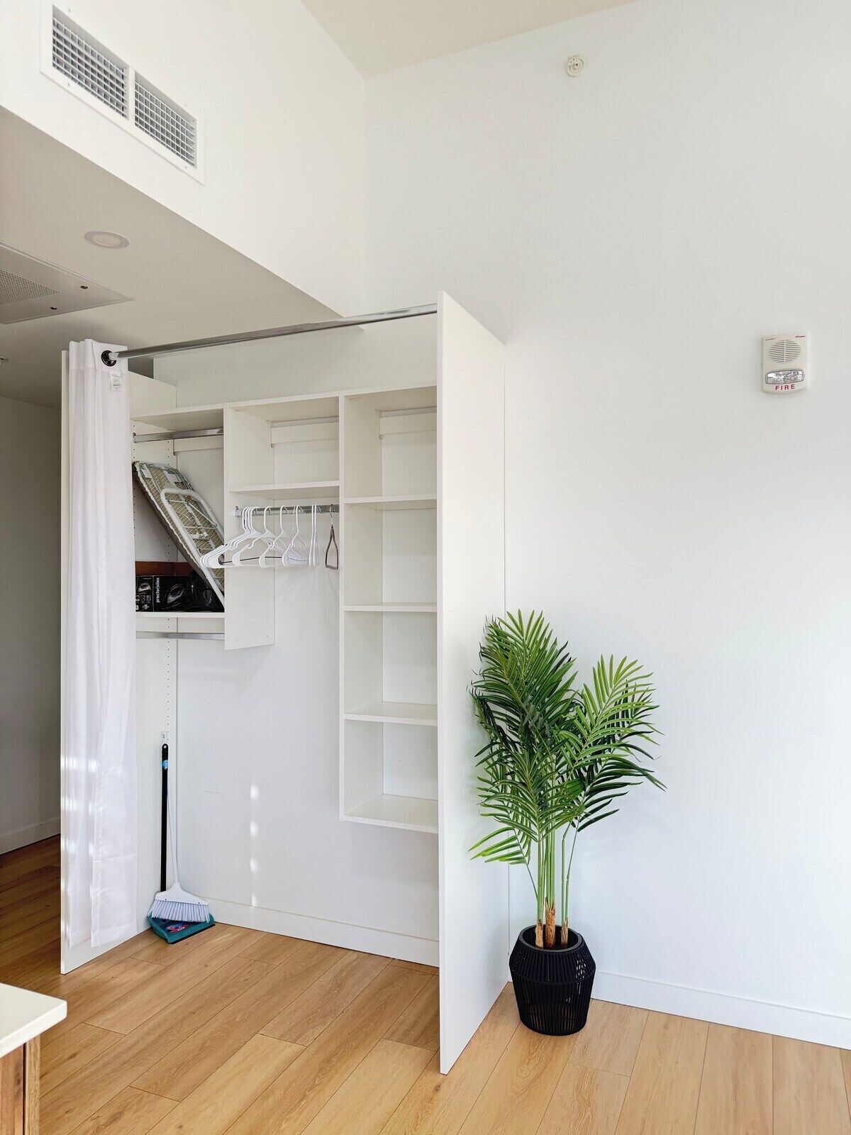 A room with a white closet, a potted plant, and a fire alarm on the wall.