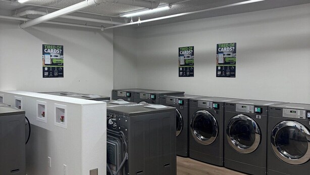 A laundry facility with multiple washing machines and dryers, fluorescent lighting, and signs on the walls. - Executive Studio Near Rogers Place” (Edmonton)