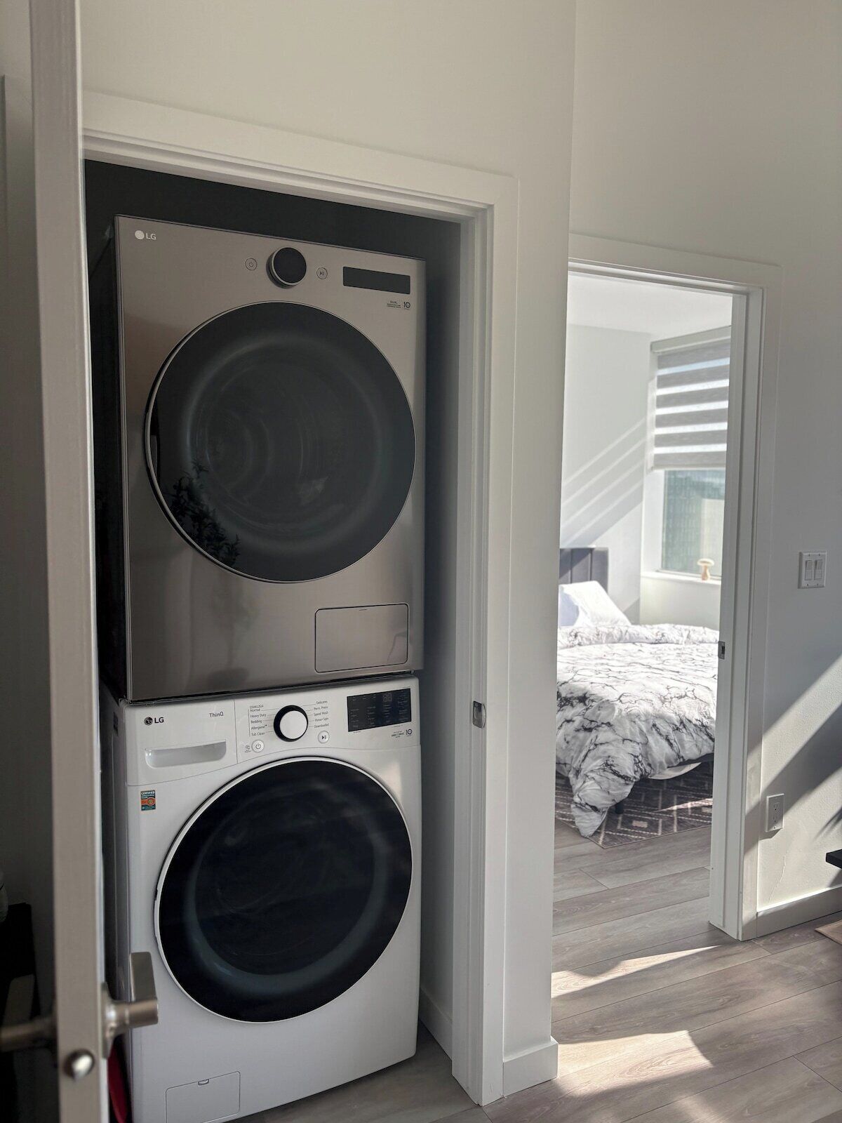 A compact laundry room with a stacked washer and dryer, a visible control panel, and a bedroom with a bed and a window.