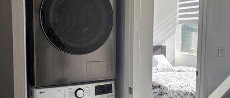 Un lavadero pequeño con lavadora y secadora apiladas, un panel de control visible y un dormitorio con cama y ventana.
