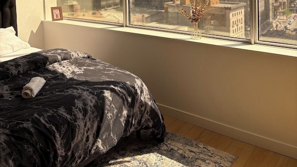 A bedroom with a large window, a bed, and a view of city buildings. - small cozy studio apartment (Edmonton)