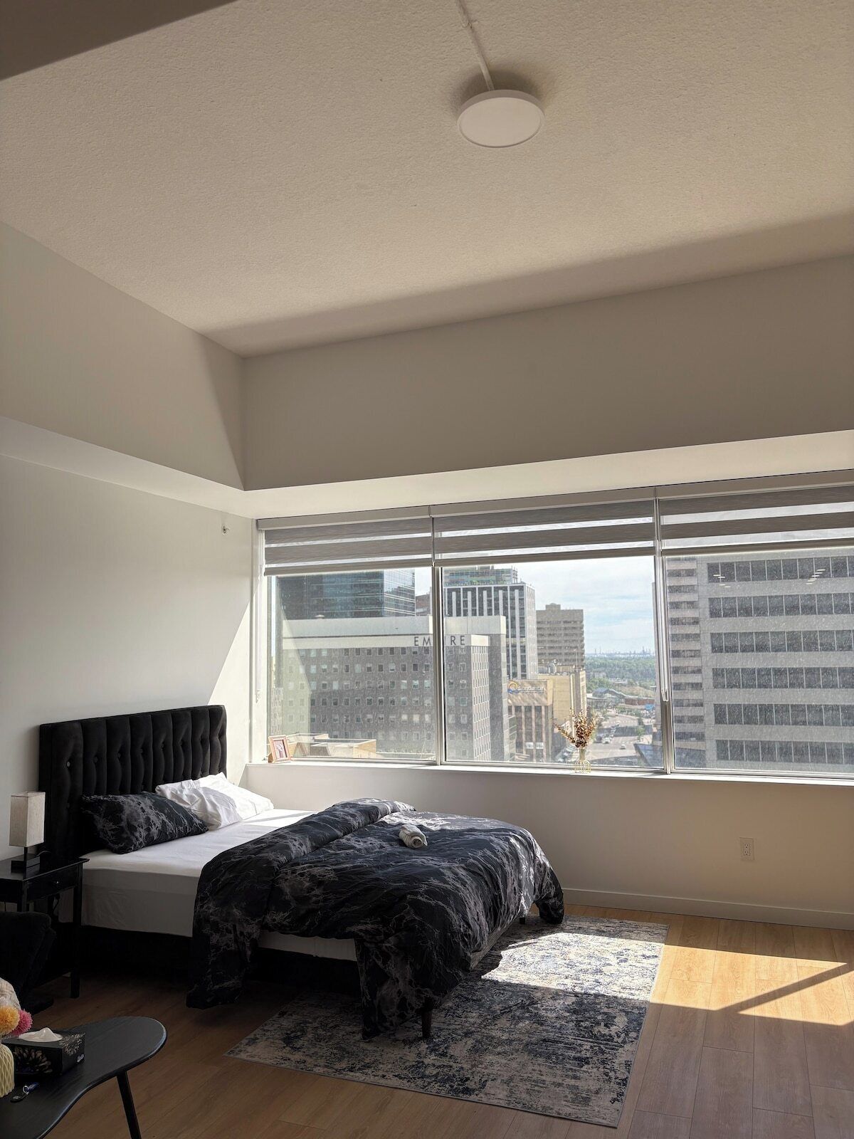 A modern bedroom with a large window, a bed with a black headboard, a bedside table, and a view of city buildings.
