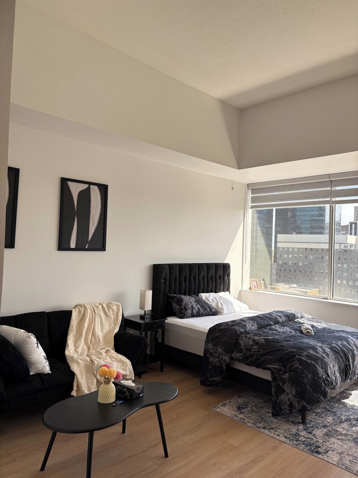 Une chambre moderne avec un lit, un canapé noir, une petite table avec un vase et une fenêtre donnant sur la ville.