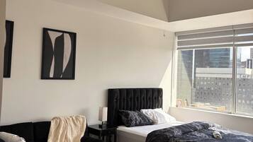 Une chambre moderne avec un lit, un canapé noir, une petite table avec un vase et une fenêtre donnant sur la ville.