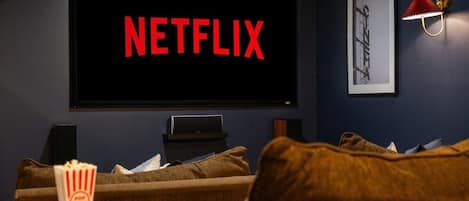 Et mørkt rum med et stort skærm der viser 'NETFLIX' i røde bogstaver, et billede i ramme på væggen og en popcornbeholder på et lille bord.