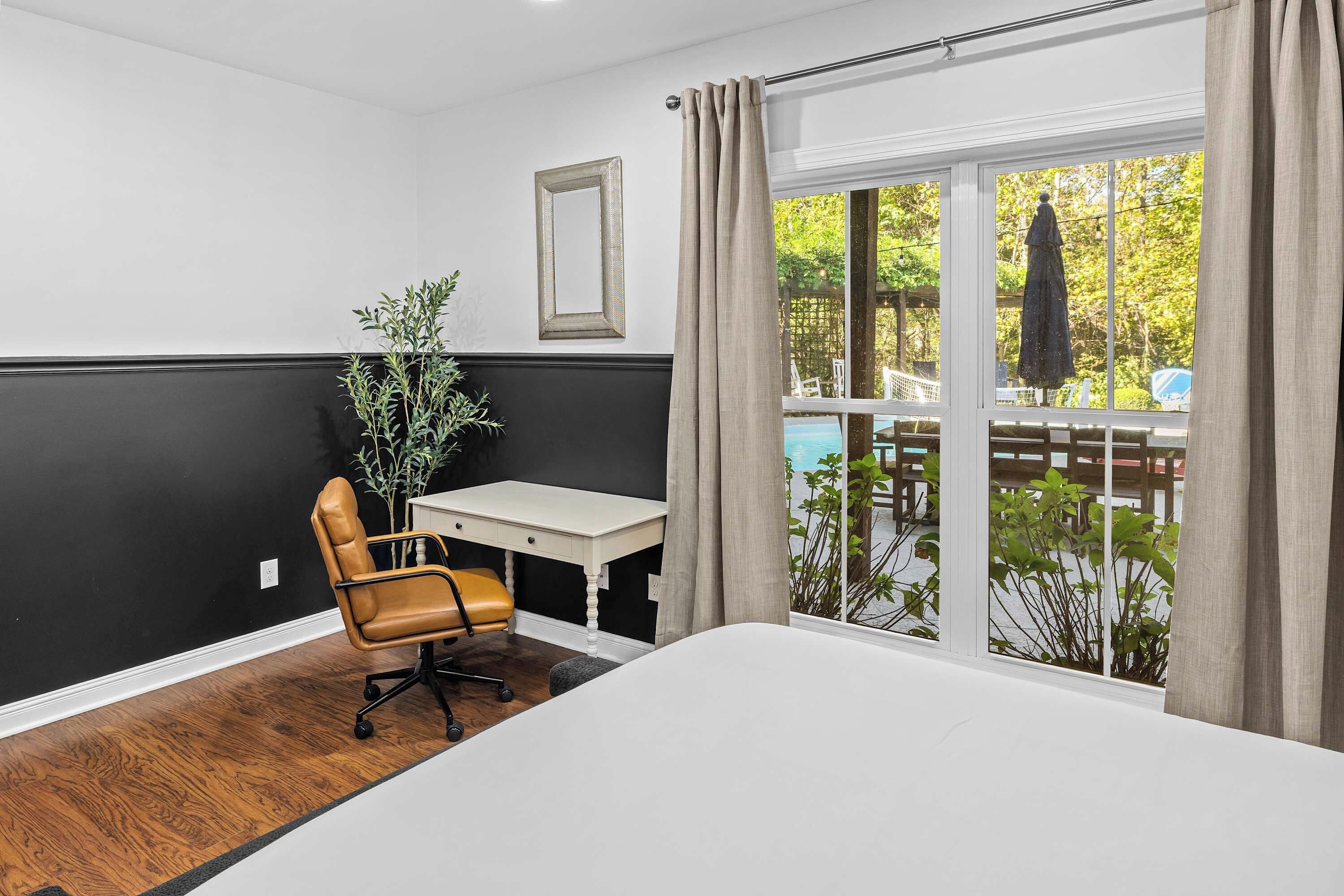 A hotel room with a bed, a desk, a chair, a potted plant, and a view of an outdoor pool area.