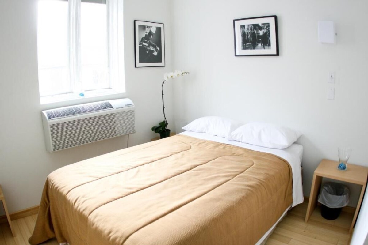 A bedroom with a bed, a window with an air conditioner, and two framed pictures on the wall.