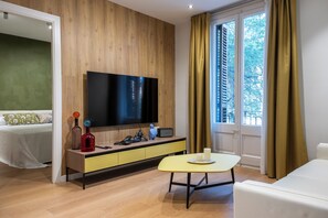 Ein modernes Hotelzimmer mit einer Wand aus Holzpaneelen, einem Flachbildfernseher, einem gelben Konsolentisch, einem Bett mit gemusterten Kissen und einem Vorhangfenster.
