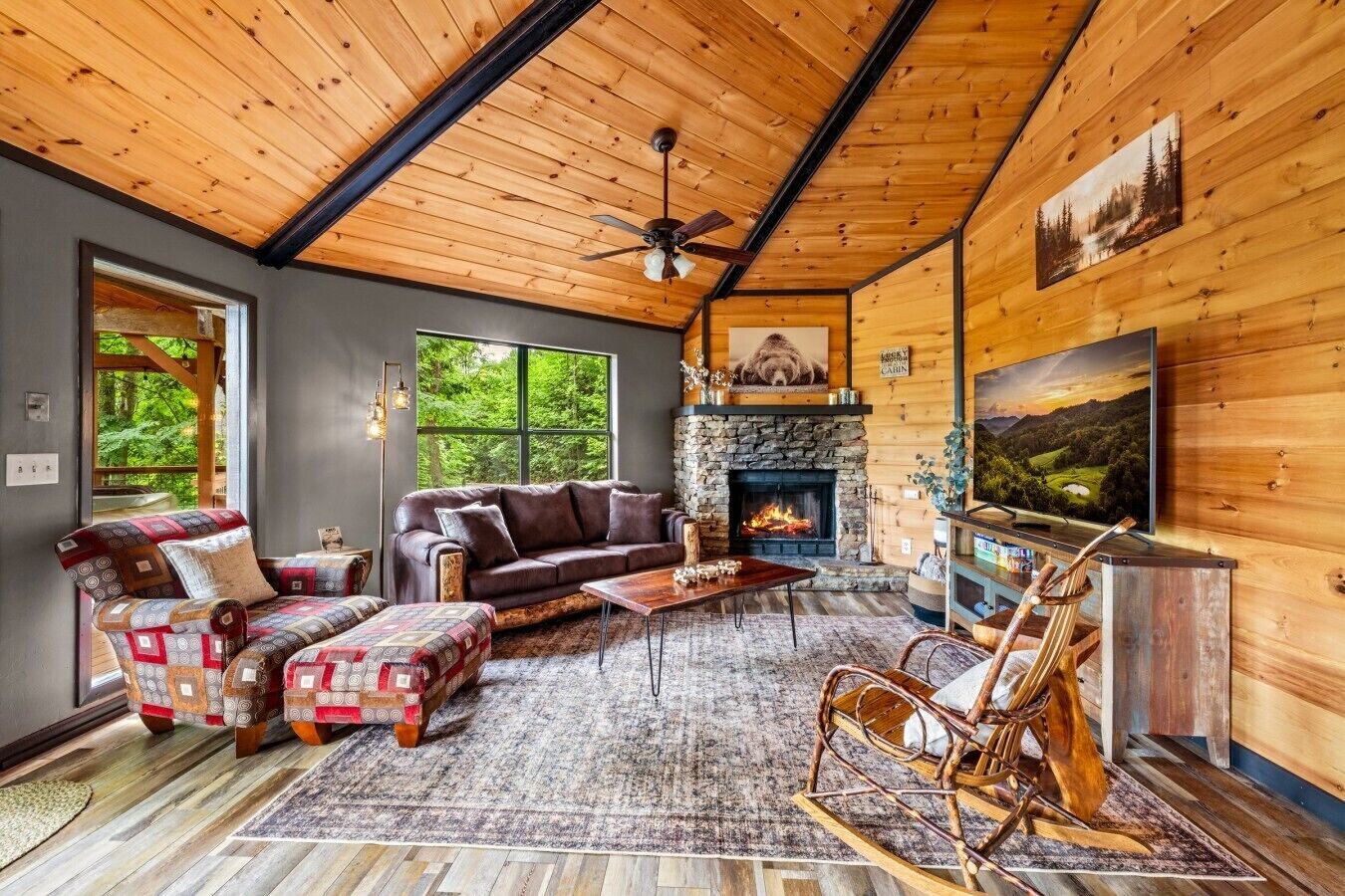 Interior acogedor de una cabaña con chimenea de piedra, un ventanal grande y techo rústico de madera.