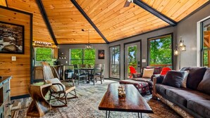 A spacious living area with a wooden ceiling, a sofa set, a dining table with chairs, and a large window providing a view of the outdoors. - Ember Hollow | Hot Tub • Near Gatlinburg • Pets (Gatlinburg)