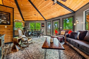 A spacious living area with a wooden ceiling, a sofa set, a dining table with chairs, and a large window providing a view of the outdoors. - Ember Hollow | Hot Tub • Near Gatlinburg • Pets (Gatlinburg)