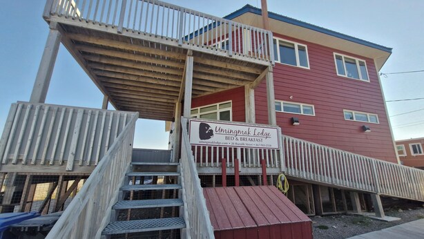 A two-story building with a sign for Umingmak Lodge, featuring a wooden staircase and a red exterior. - Umingmak Lodge Bed And Breakfast (Cambridge Bay)