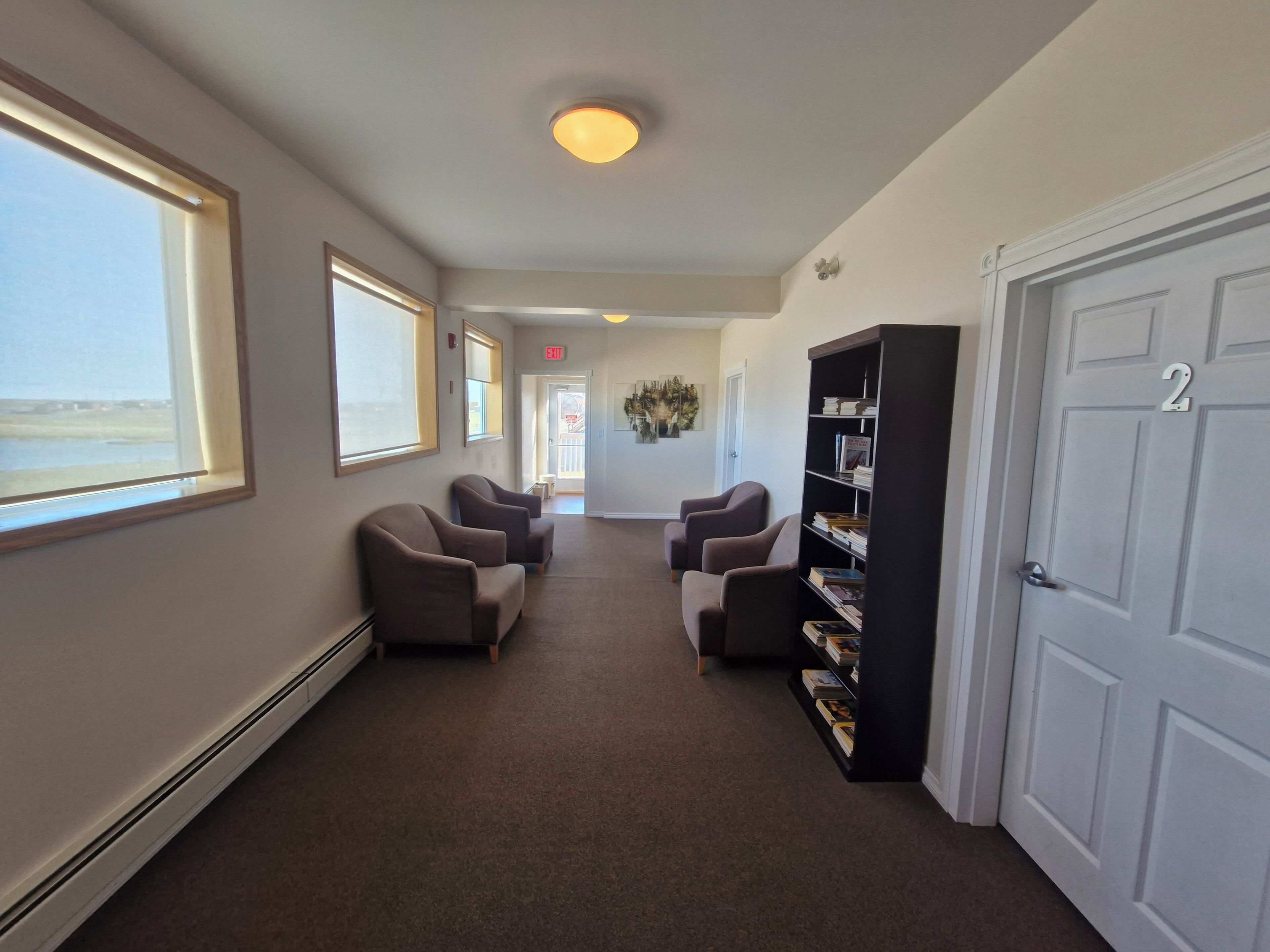 A hallway with a carpeted floor, a row of armchairs, a bookshelf, and a door with a number.