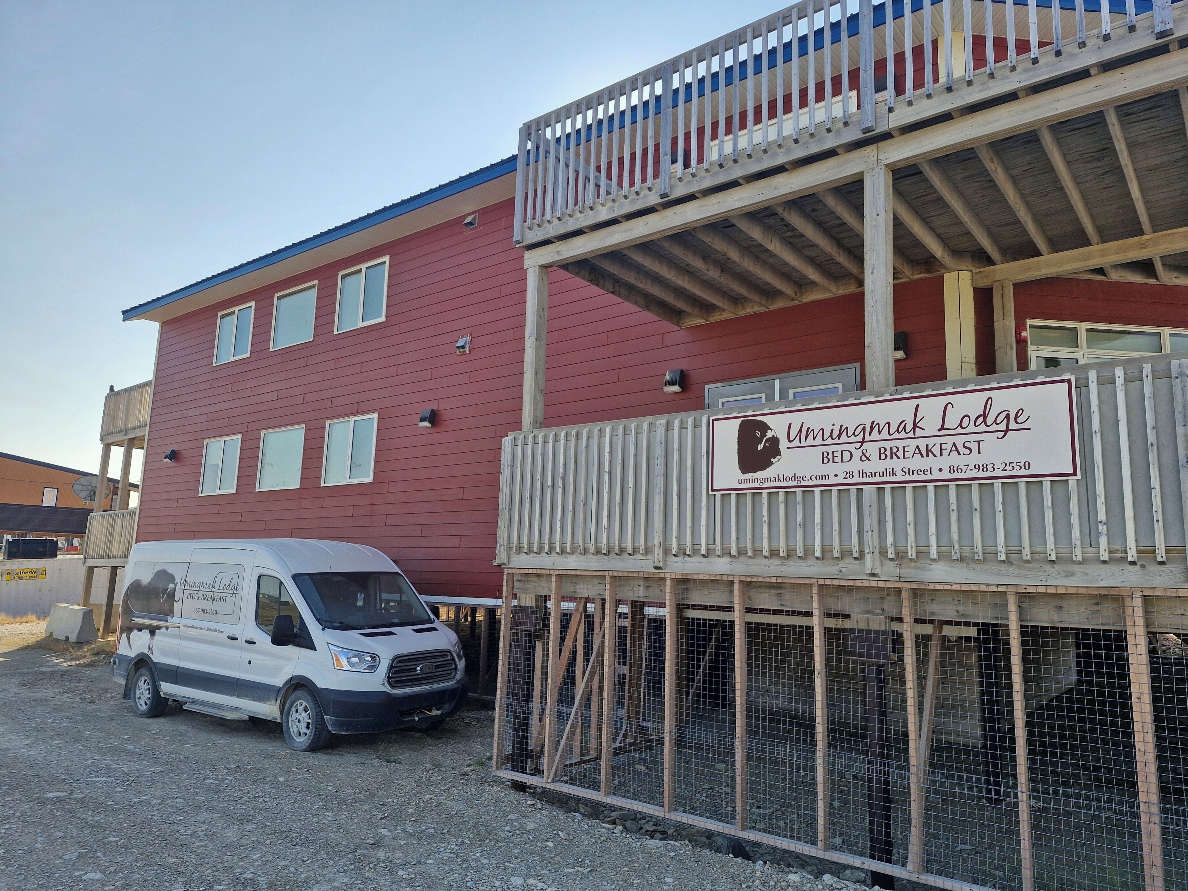 A two-story building with a sign for Umingmak Lodge Bed & Breakfast, a white van parked in front, and a wooden fence with a railing.