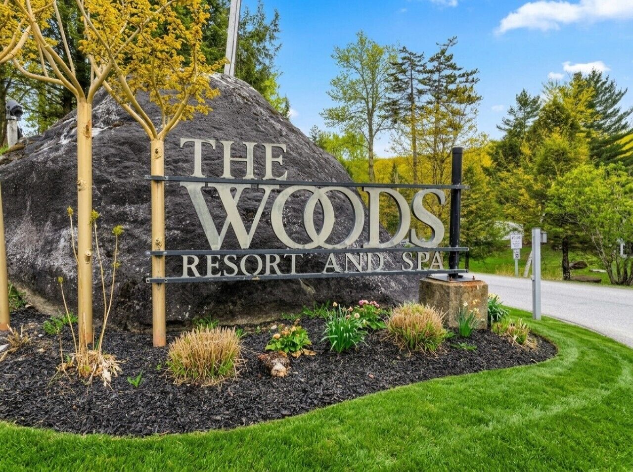 Uma rocha grande com a placa 'The Woods Resort and Spa', rodeada por um jardim paisagístico com diversas plantas.