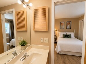 Une chambre d’hôtel avec une salle de bain comprenant un lavabo, un miroir et une plante en pot. On y trouve un lit avec un oreiller vert et un couvre-lit blanc.