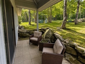 An outdoor patio area with two wicker chairs and a glass-top table, surrounded by a stone wall and a grassy lawn with trees. - 7 minutes from the Killington Base Lodge and seconds away from the best restaurants and nightlife! (Killington)