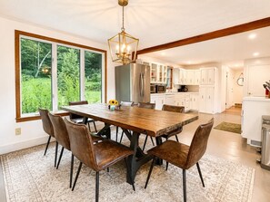 A dining area with a wooden table and chairs, a chandelier, and a view of a kitchen and garden. - Near Killington & Woodstock | Game Room, Fireplace (Bridgewater)