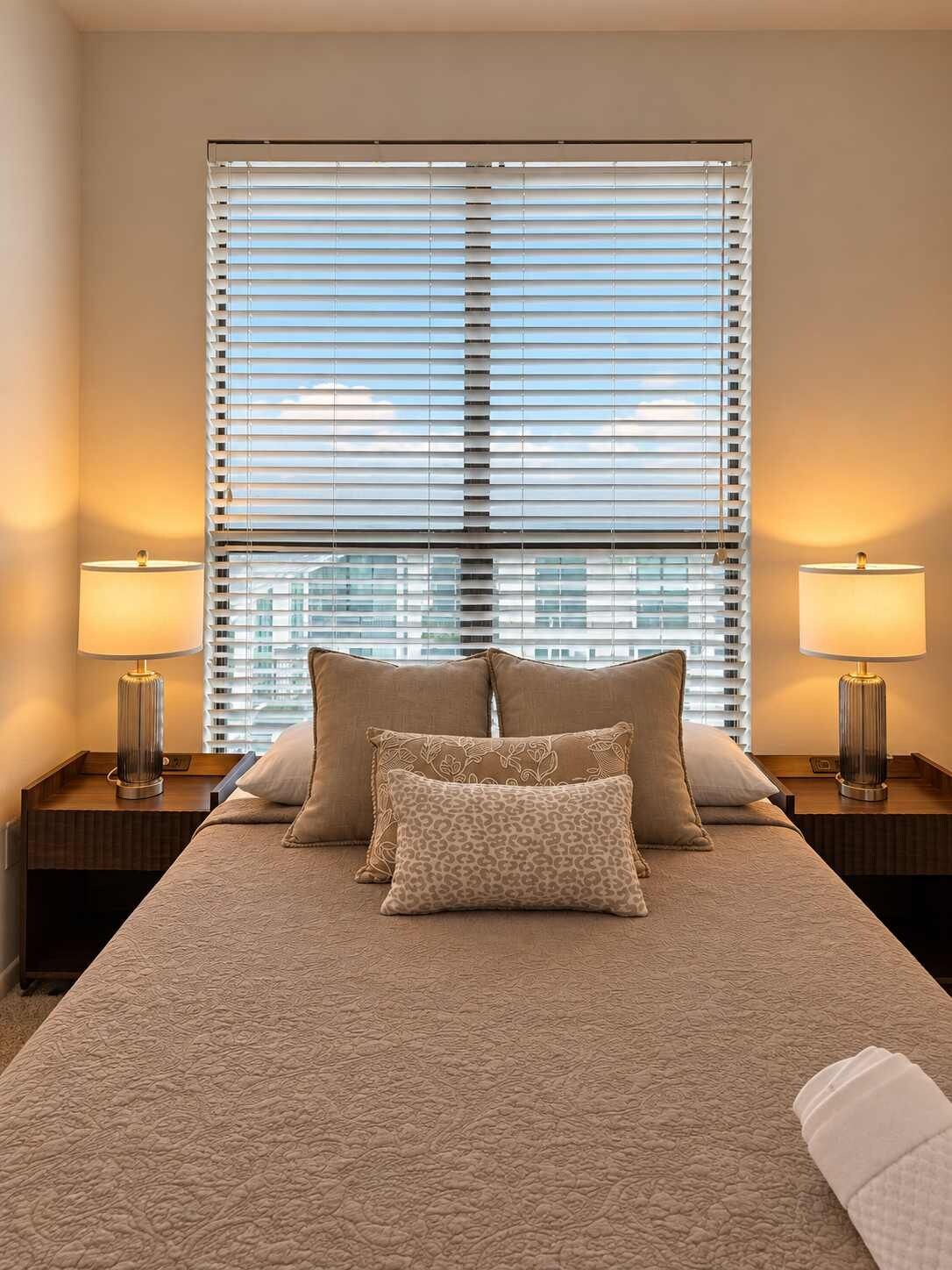 A hotel room with a large bed, two bedside lamps, and a window with blinds.